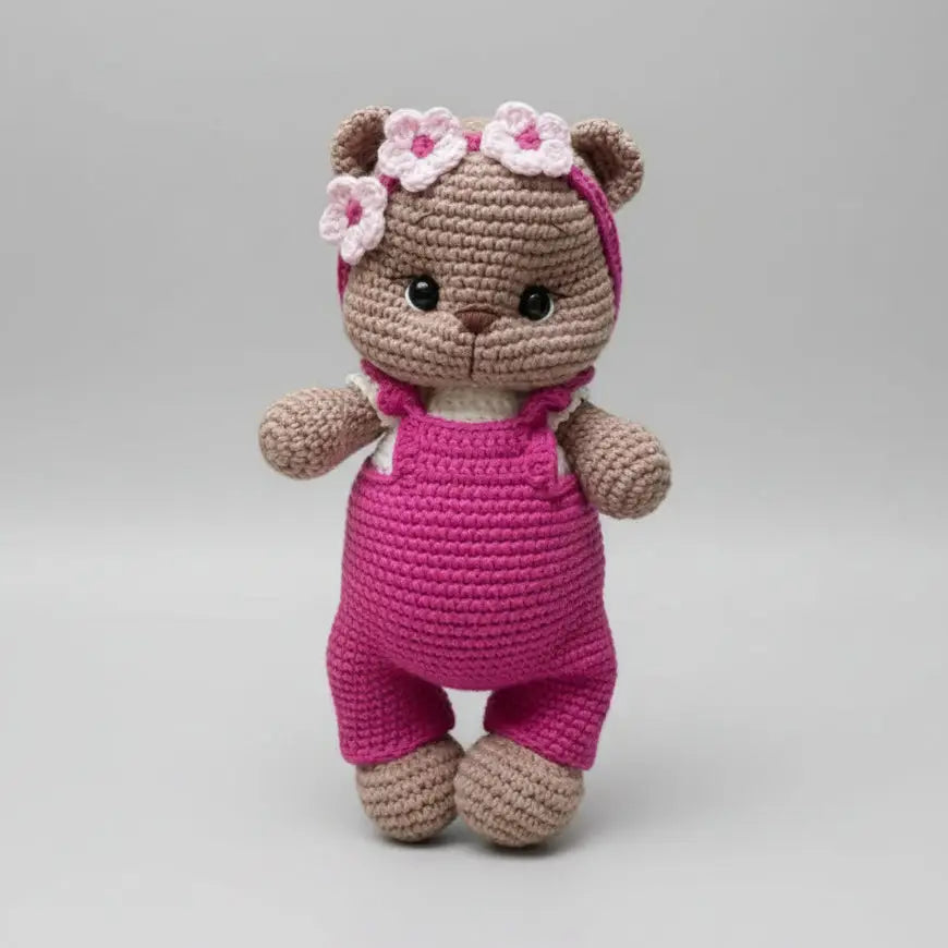Crocheted teddy bear in pink overalls with flower crown, standing on gray background