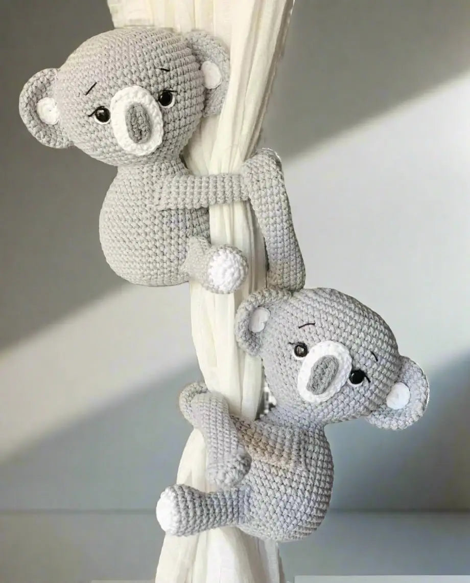 Two crocheted gray koala toys hugging a white curtain or drape