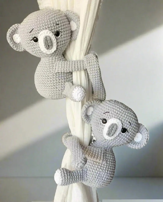 Two crocheted gray koala toys hugging a white curtain or drape