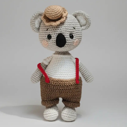 Crocheted koala wearing hat, brown pants, and red suspenders on gray background