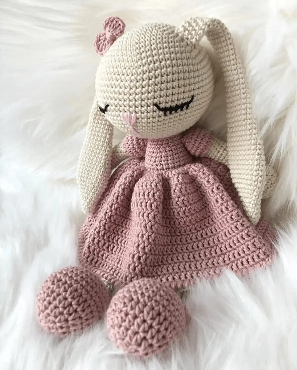 Crocheted bunny doll in pink dress, resting on fluffy white surface with closed eyes