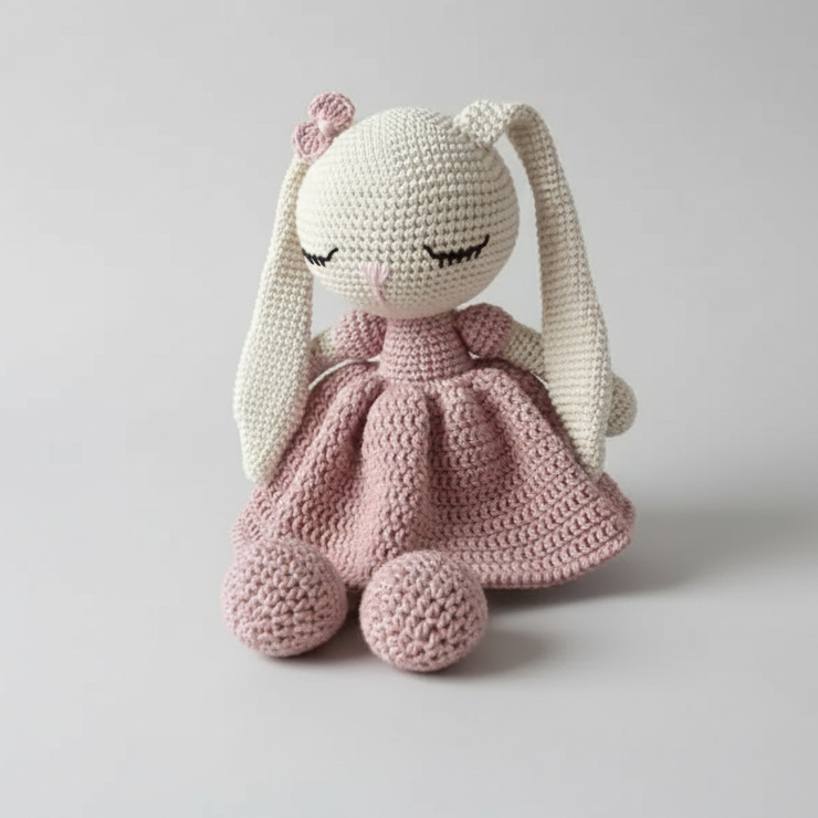 Crocheted bunny doll in pink dress, resting on fluffy white surface with closed eyes
