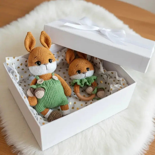 Two crocheted bunny dolls in green and orange nestled in a white gift box with gold heart pattern.