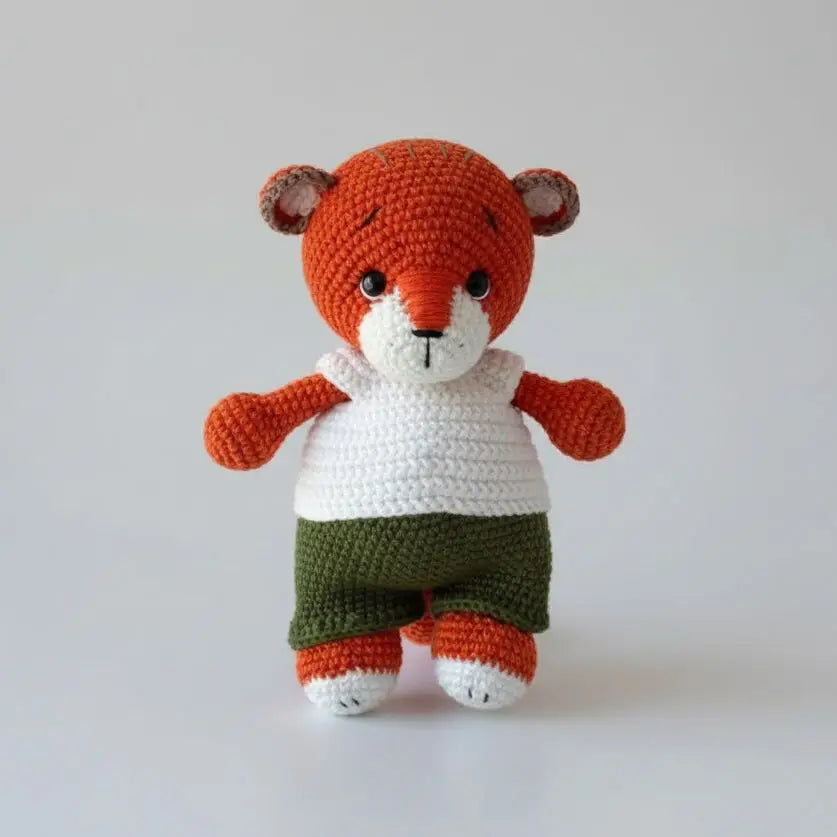 Crocheted orange teddy bear with white shirt and green shorts, standing upright