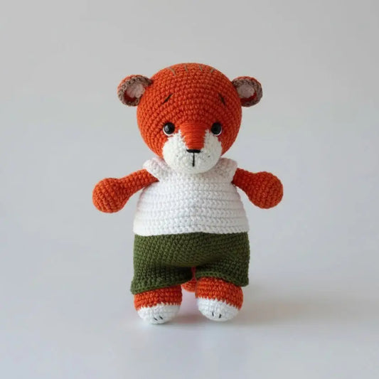 Crocheted orange teddy bear with white shirt and green shorts, standing upright