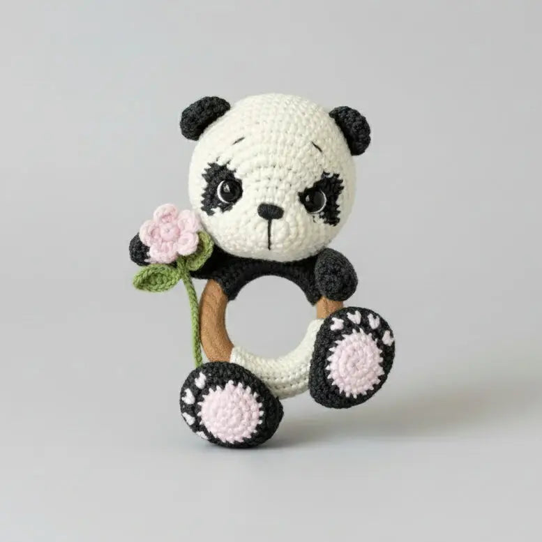 Crocheted panda bear holding a pink flower, sitting against a gray background.