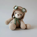 Crocheted teddy bear in aviator hat and scarf, sitting against neutral background