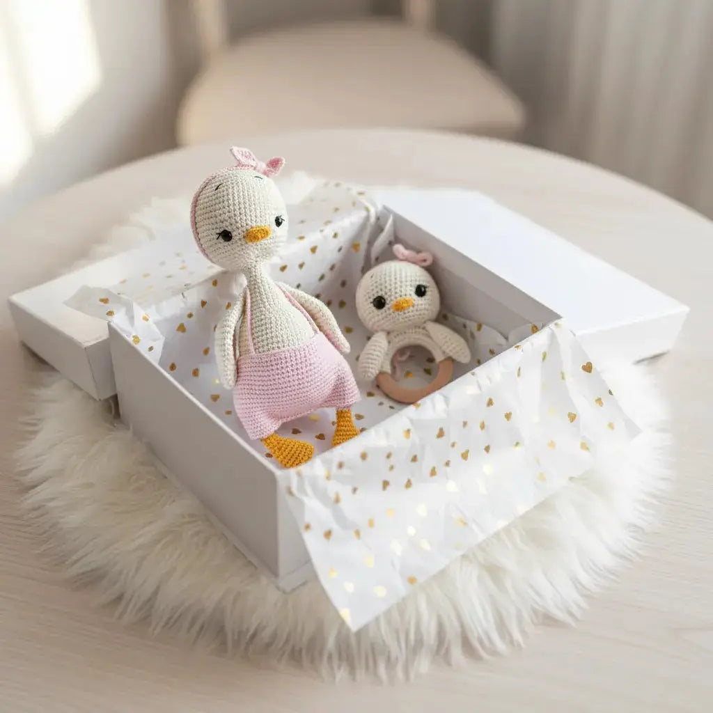 Two crocheted penguin dolls, one larger with a pink dress and bow, the other smaller with a teething ring, nestled in a white box with gold heart-patterned tissue paper on a fluffy white surface.