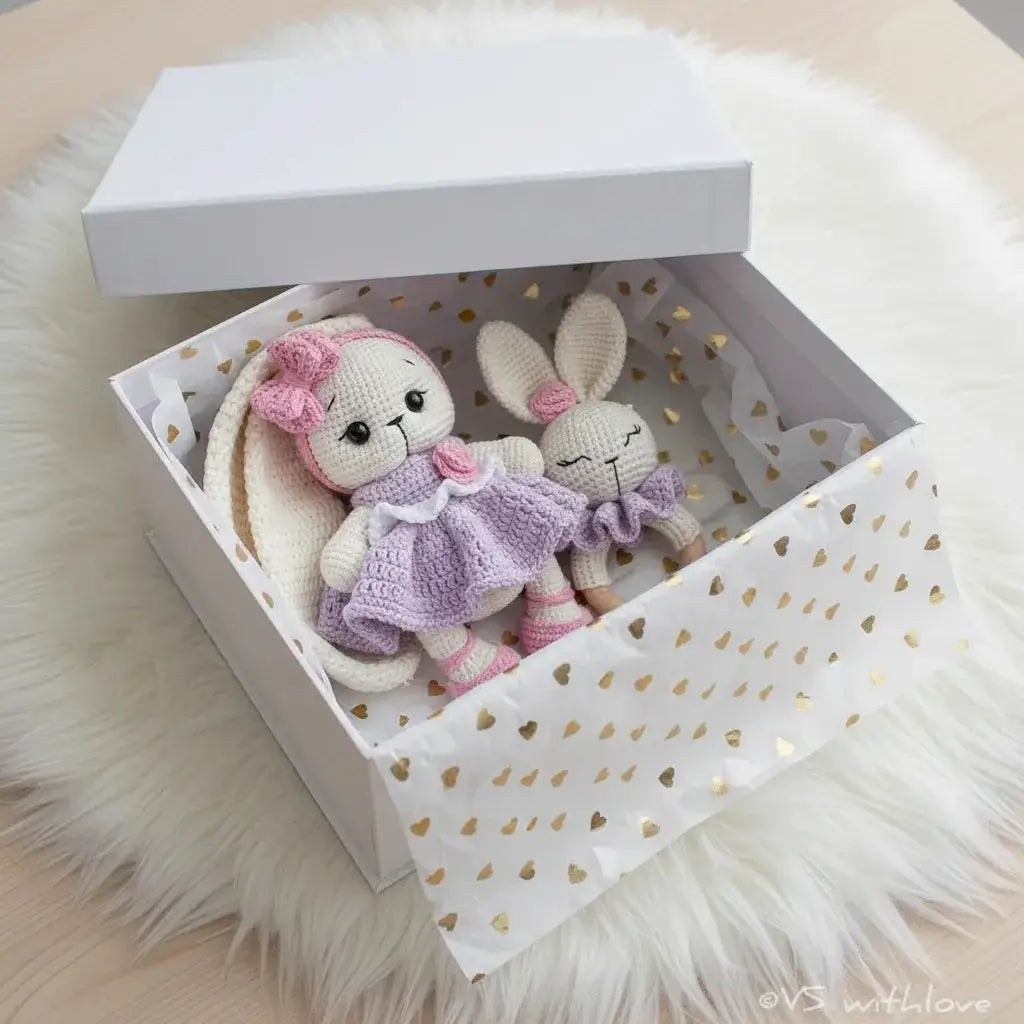 Two crocheted bunny dolls in lavender and pink inside a heart-patterned gift box on fluffy white fabric
