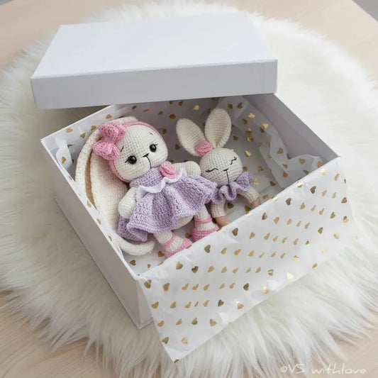 Two crocheted bunny dolls in lavender and pink inside a heart-patterned gift box on fluffy white fabric