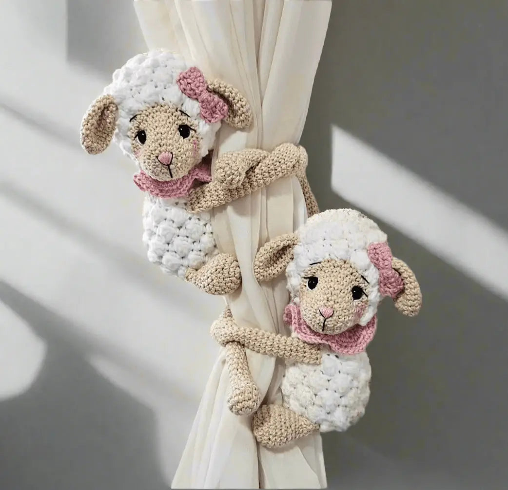 Two crocheted lamb curtain tie-backs with pink bows, hugging a white curtain.