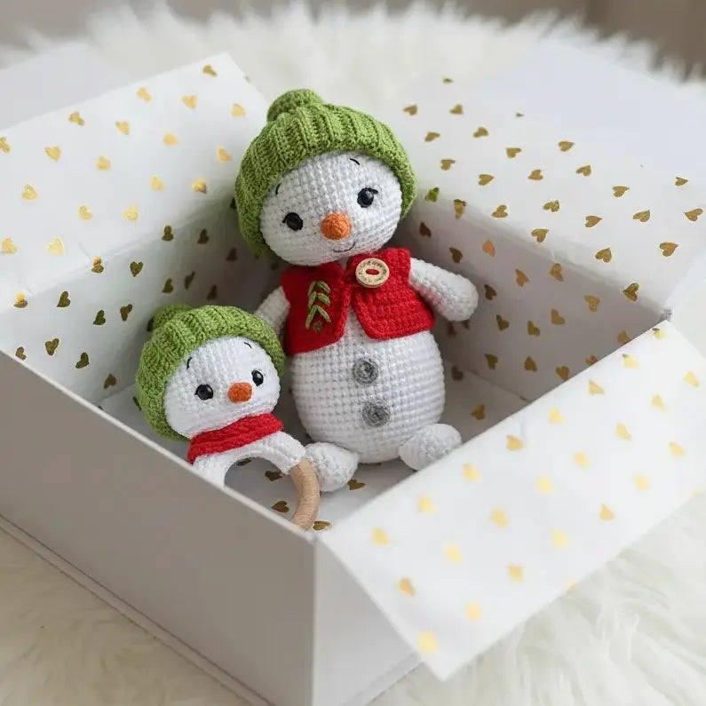 Two small crochet snowman toys in a white box with gold heart pattern on a fluffy white surface.