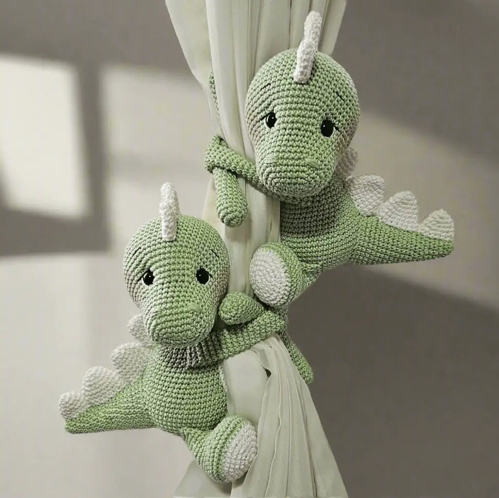 Two crocheted green dinosaur plushies hugging a white curtain, with soft details and cute expressions.