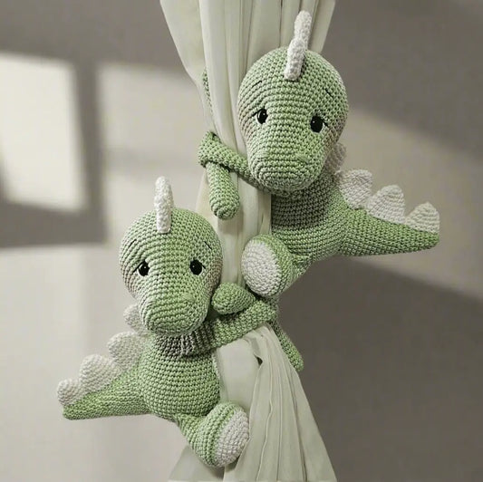 Two crocheted green dinosaur plushies hugging a white curtain, with soft details and cute expressions.