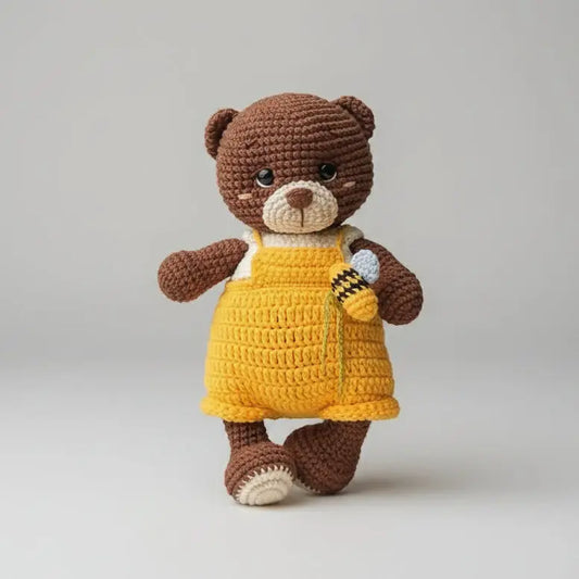 Crocheted brown teddy bear wearing yellow overalls, standing on a gray background