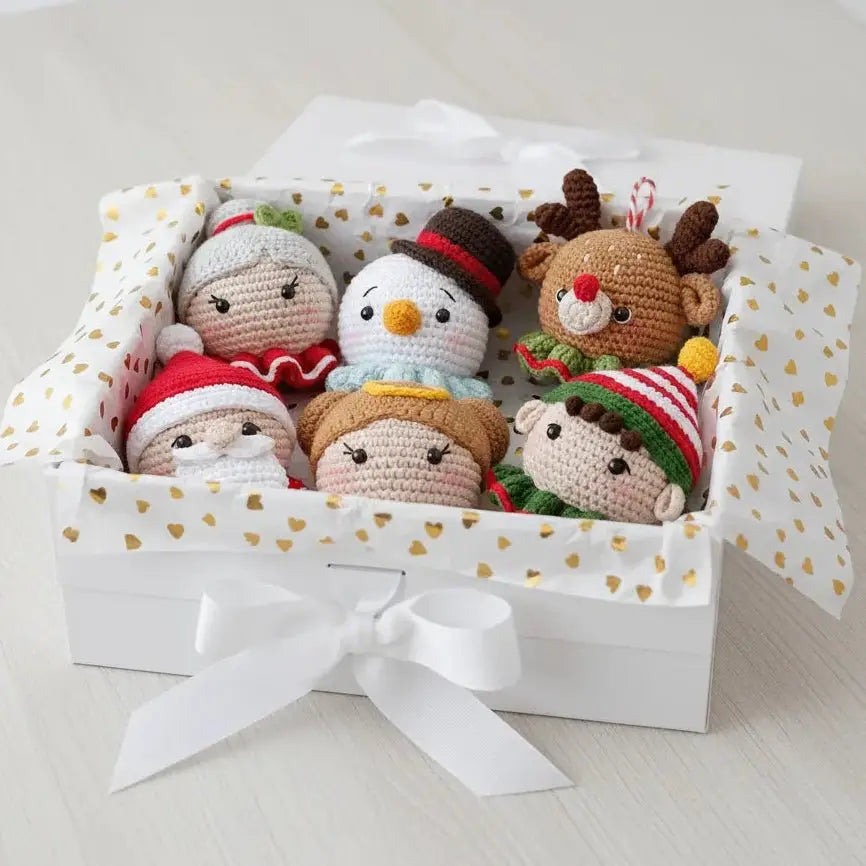 Six crocheted holiday characters nestled in a white gift box with gold heart pattern and white bow