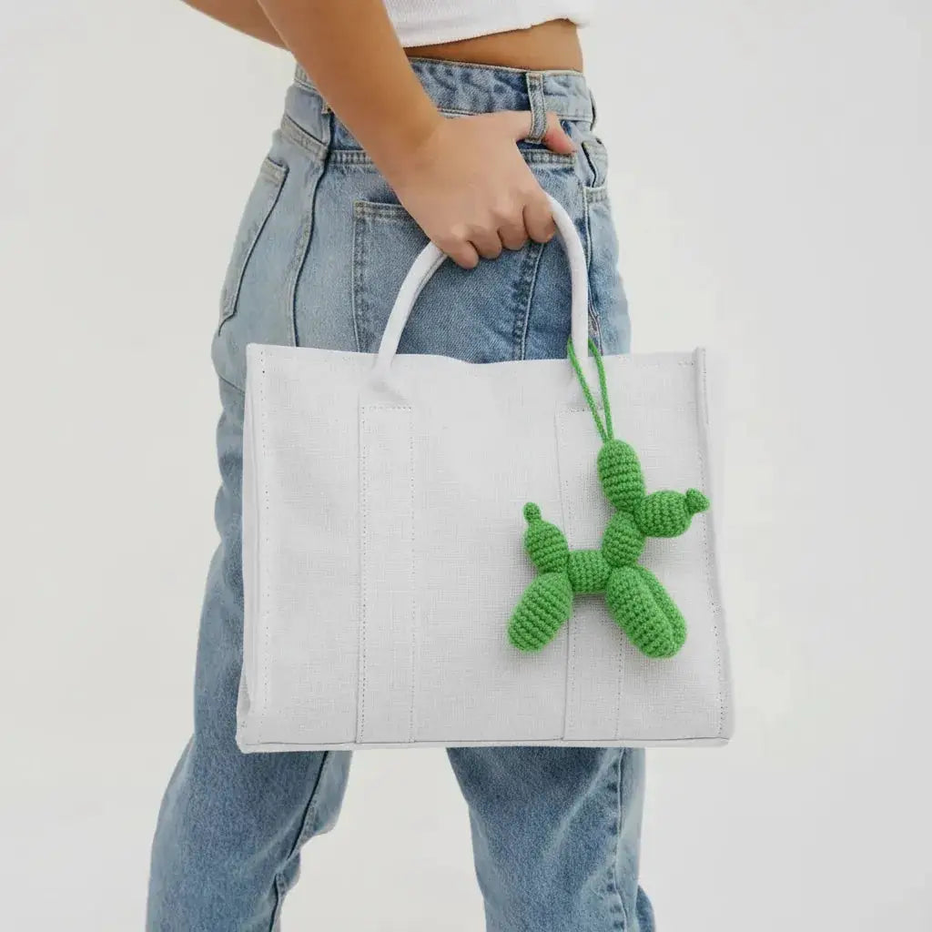 White tote bag with green crocheted balloon dog charm, held by person in denim jeans