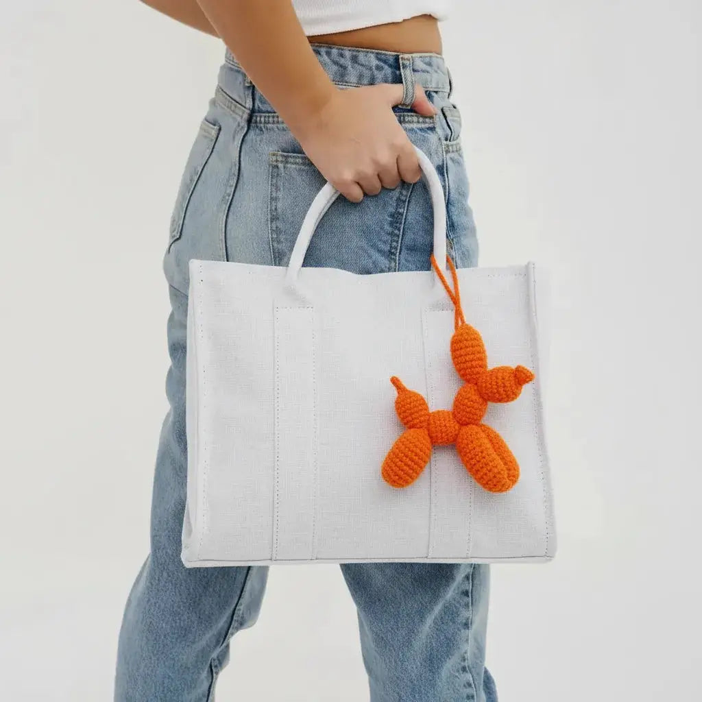 White tote bag with orange crocheted balloon dog charm, carried by someone in light blue jeans and a white crop top.