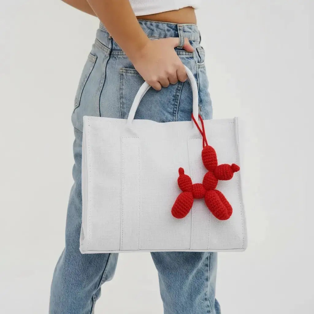White tote bag with red crocheted balloon dog charm, held against light blue denim jeans.