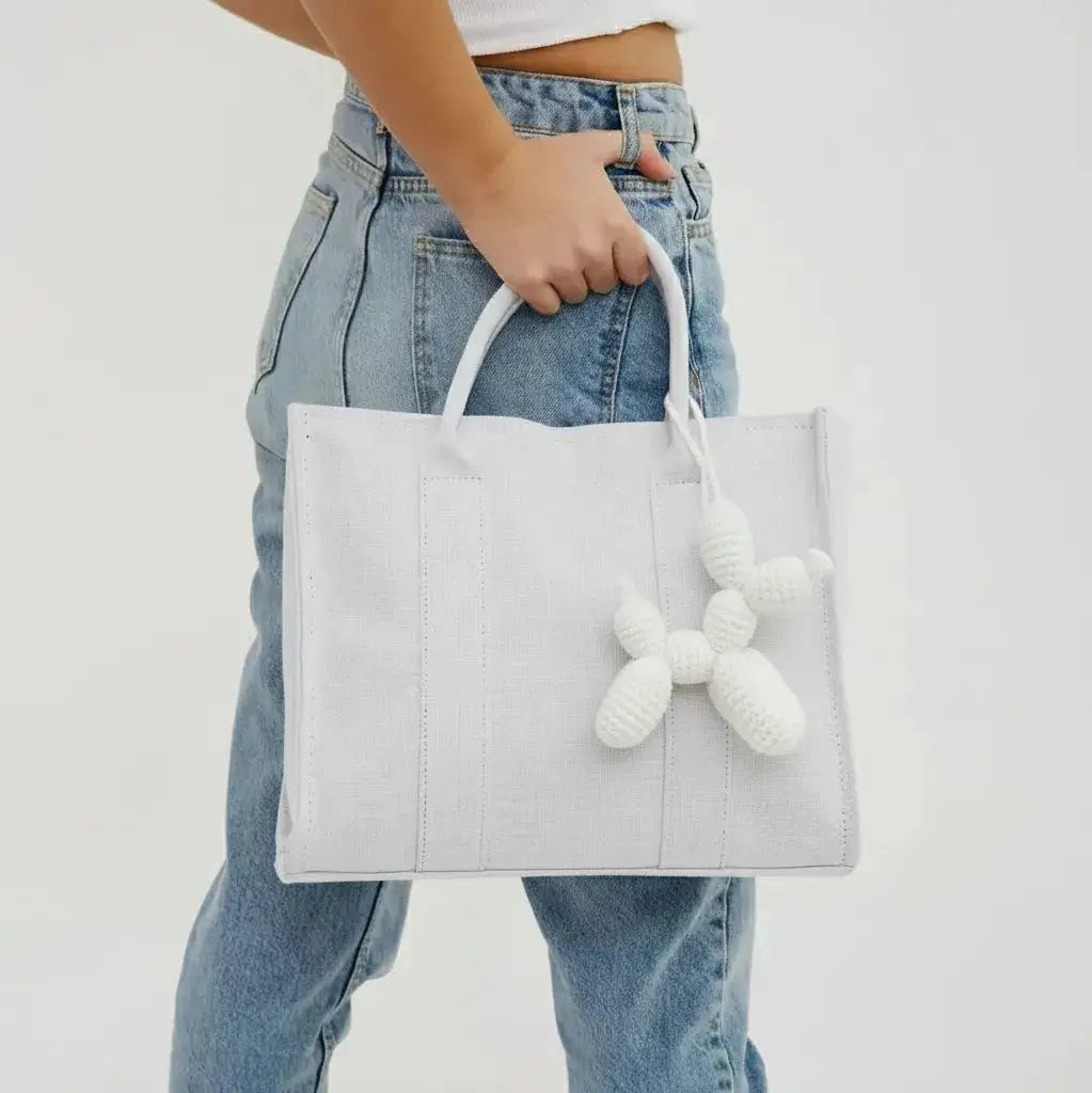 White structured tote bag with knitted white charm, held by hand against light blue denim jeans.