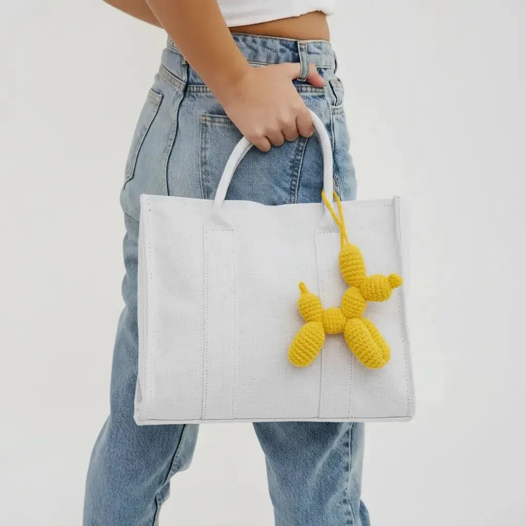 White tote bag with yellow crocheted balloon dog charm, held by a hand wearing blue denim jeans.