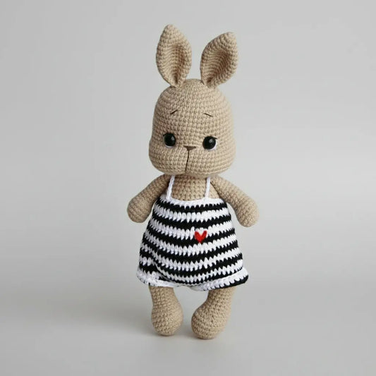 Crocheted bunny in striped dress with tiny red heart, standing against neutral background