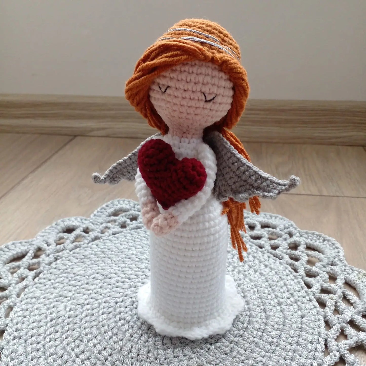 Crocheted angel figurine with red heart, orange hair, and gray wings on doily