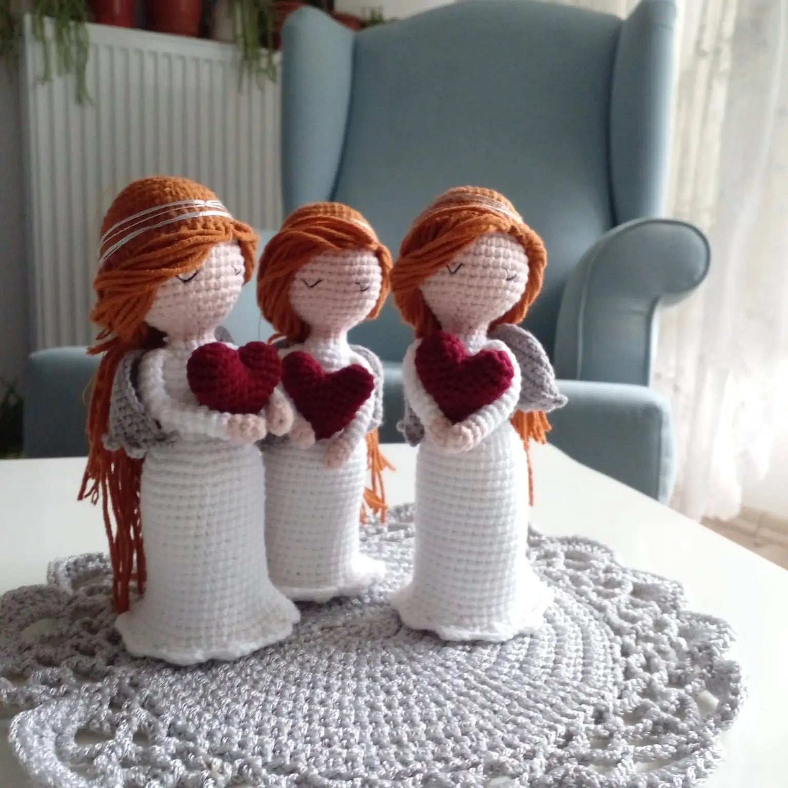 Three crocheted angel dolls with red hearts, standing on a gray doily in front of a blue chair