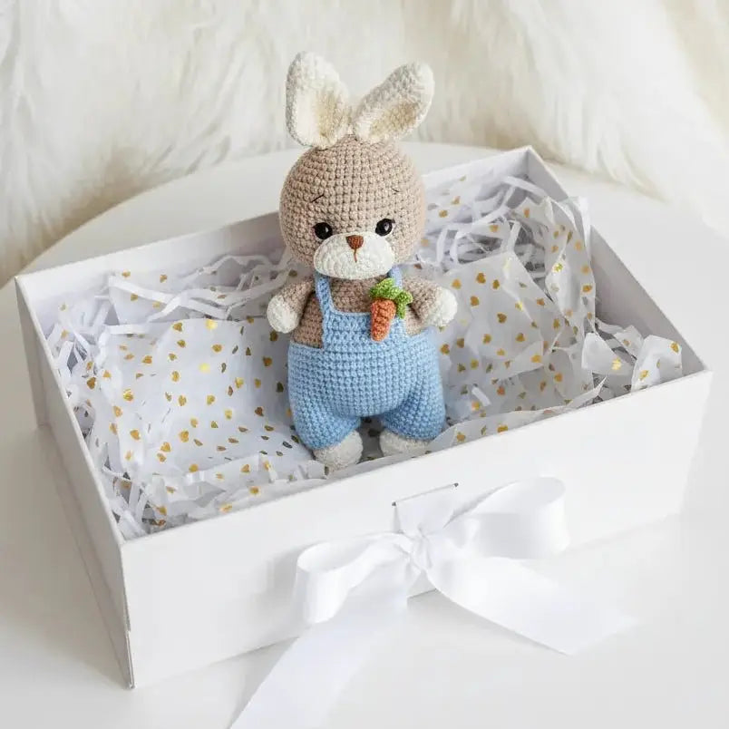 Crocheted bunny in blue overalls with carrot, nestled in a white gift box with gold dots