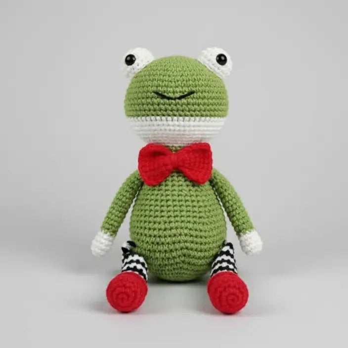 Crocheted green frog plush with red bow tie and striped socks, sitting upright