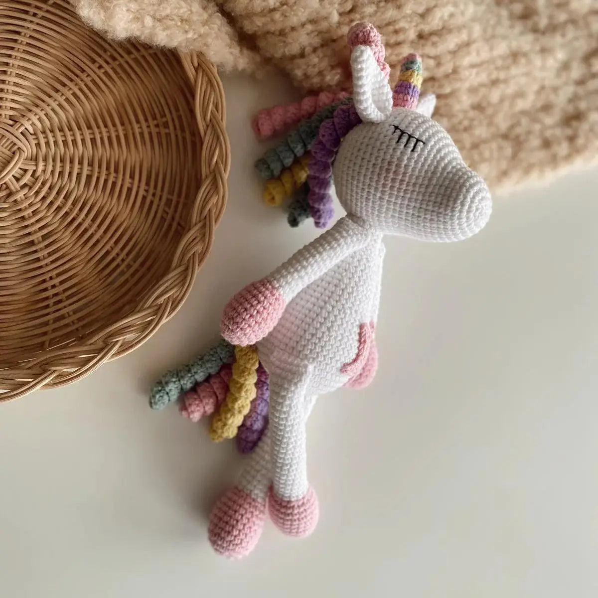 Crocheted unicorn with rainbow mane and tail, white body, resting beside woven basket