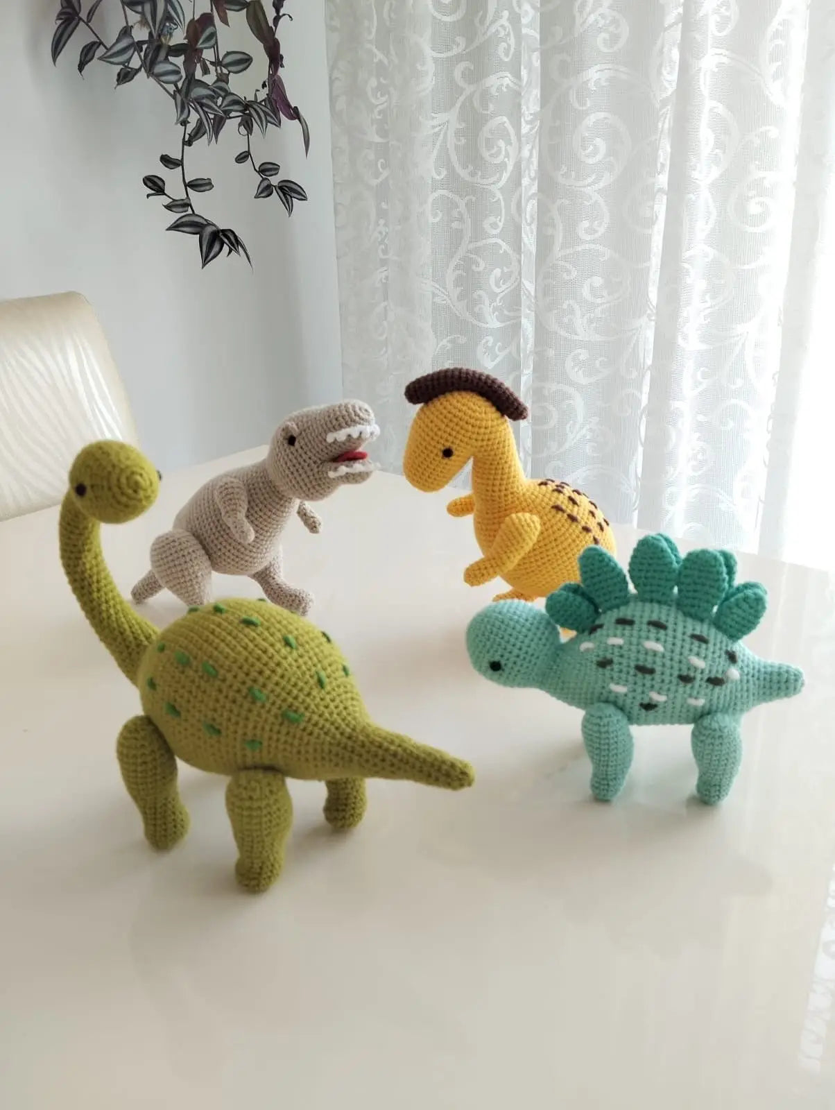 Four crocheted dinosaurs in different colors and species, standing together on a white surface