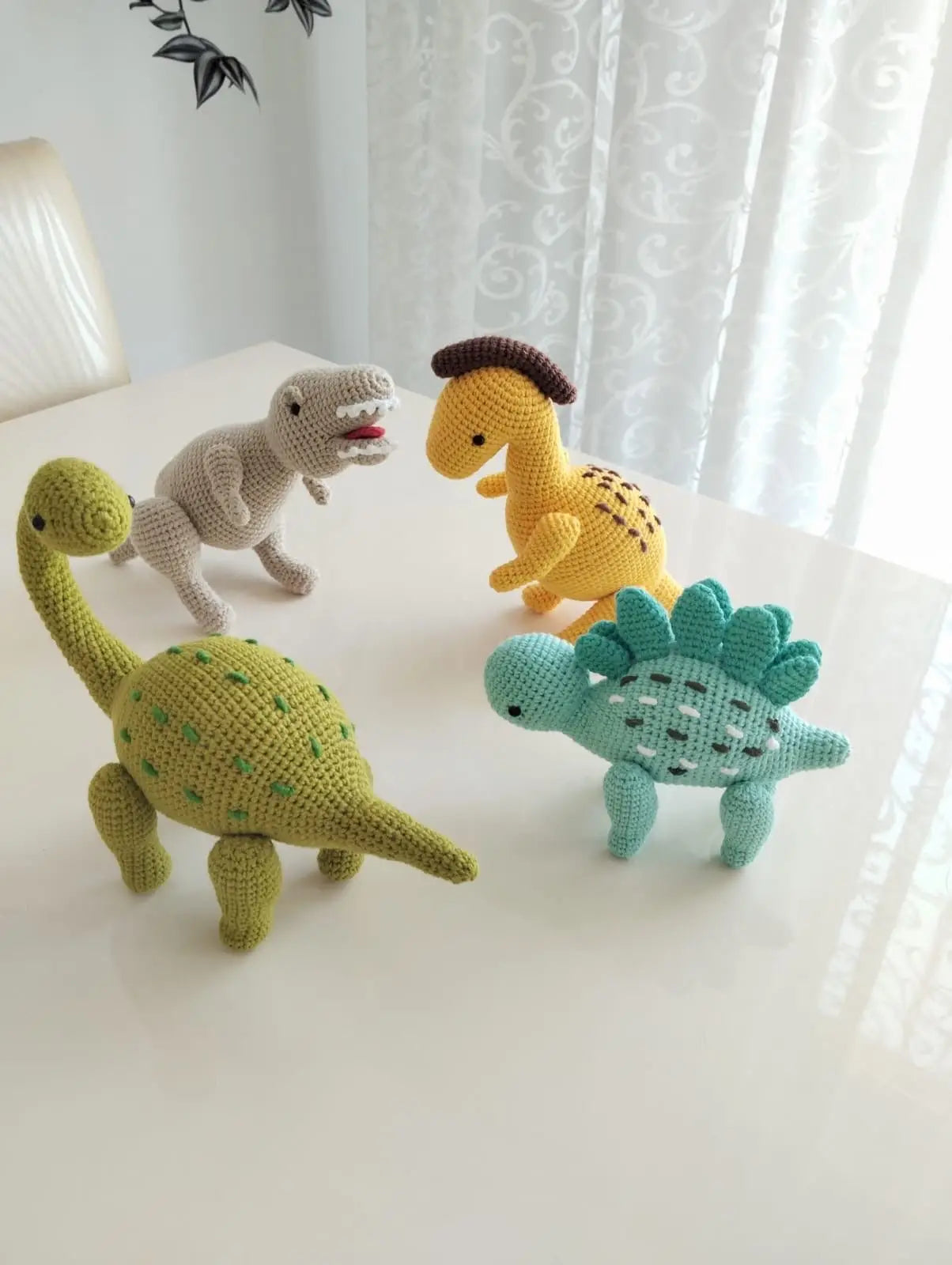 Four colorful crocheted dinosaurs in green, beige, yellow, and teal arranged on a white surface.