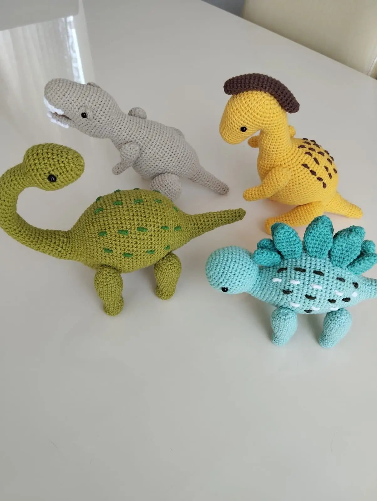 Crocheted dinosaurs in green, gray, yellow, and teal on a white surface.