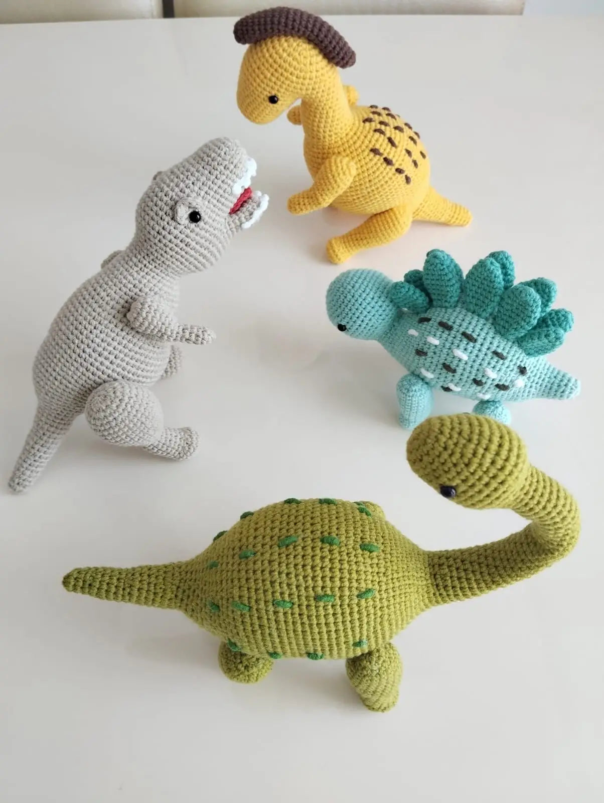Crocheted dinosaurs in yellow, green, blue, and gray, posed playfully on a white surface