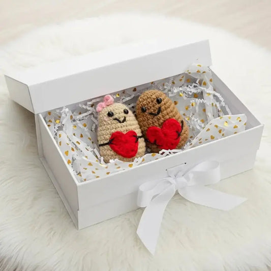 Two crocheted teddy bears with red hearts in a white gift box with gold confetti liner