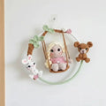 Crocheted doll on swing with bunny and teddy bear, surrounded by pastel leaves and flowers