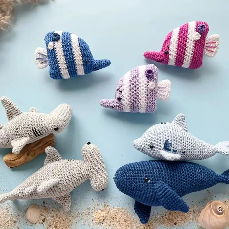 Crocheted marine animals: sharks, fish in blue, pink, and purple stripes on sandy background