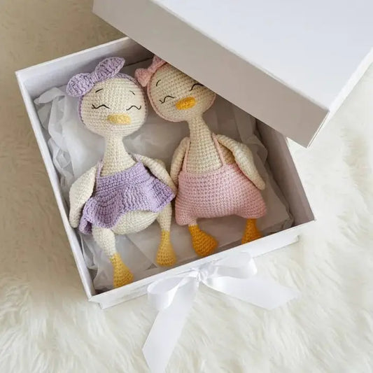 Two crocheted duck dolls in purple and pink outfits nestled in a white gift box with tissue paper