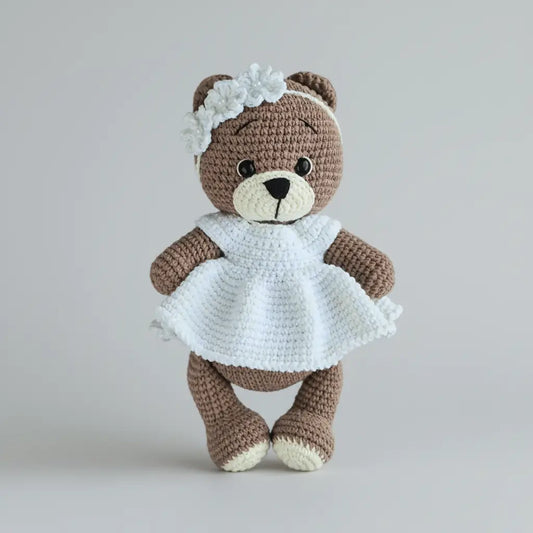 Crocheted teddy bear in white dress with flower headband, standing against gray background