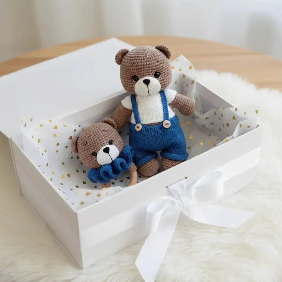 Two crocheted teddy bears in blue overalls, nestled in a white gift box with golden confetti.