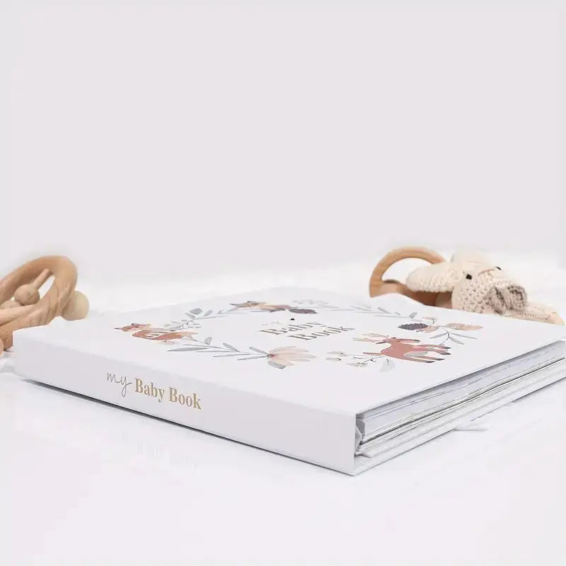 White baby book with floral design, wooden teething rings, and knit stuffed toy nearby