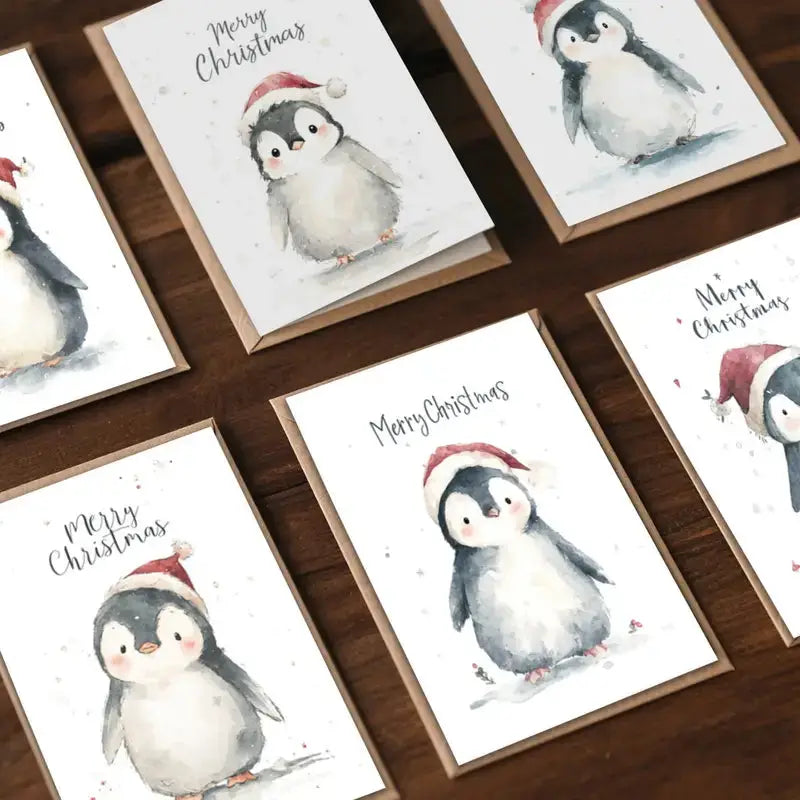 Watercolor Christmas cards featuring cute penguins in Santa hats, laid out on a wooden surface.