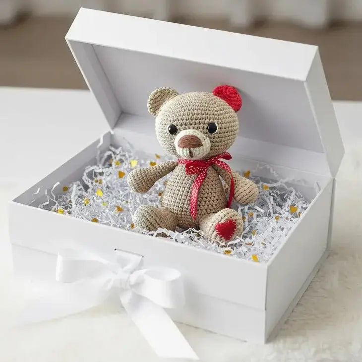 Crocheted teddy bear with red accents nestled in a white gift box with shredded paper