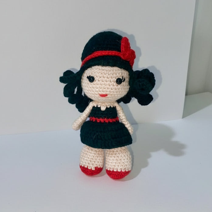 Crocheted doll with black curly hair, black dress, red hat, and red shoes, standing on a white surface.