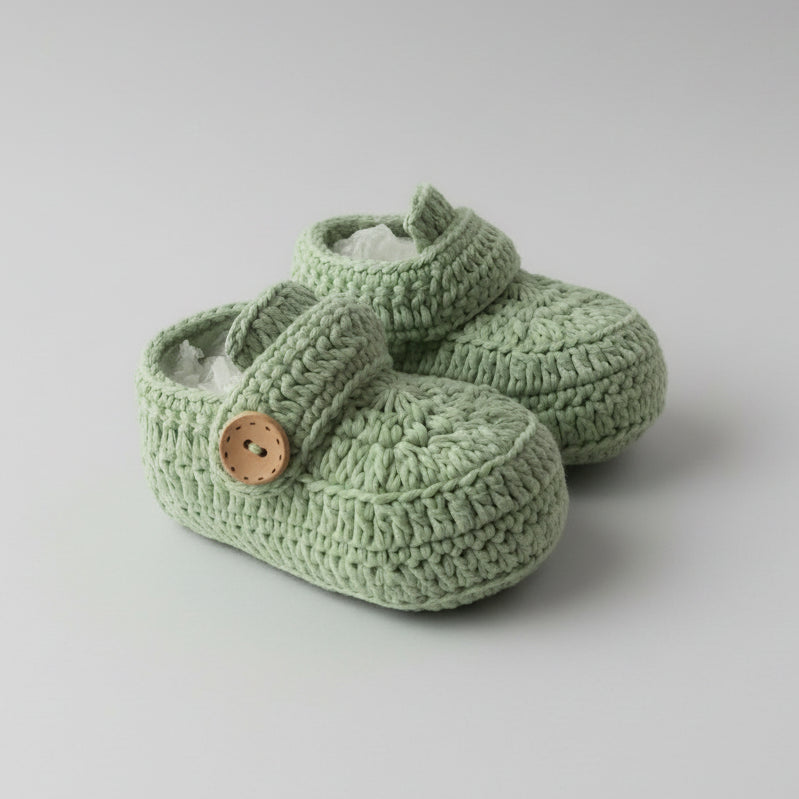 Sage green crocheted baby booties with wooden button, soft textured knit on wooden surface