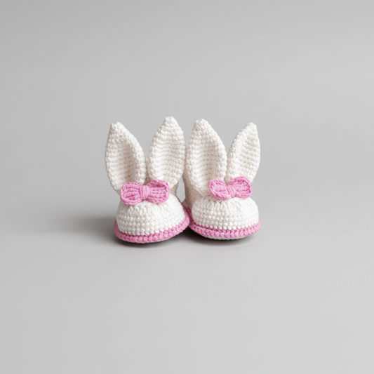 Crocheted white baby booties with pink bows, shaped like bunny ears, on wooden surface