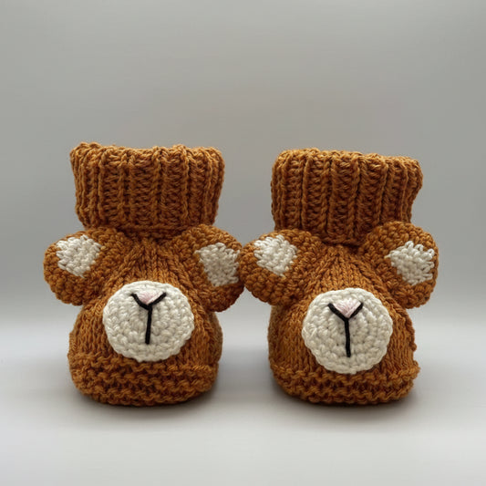 Knitted baby booties with bear faces, brown with white accents, on pink background