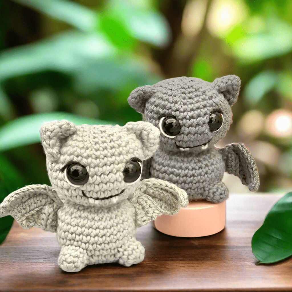 Two crocheted bat plushies, one white and one gray, sitting on a wooden surface with green leaves.