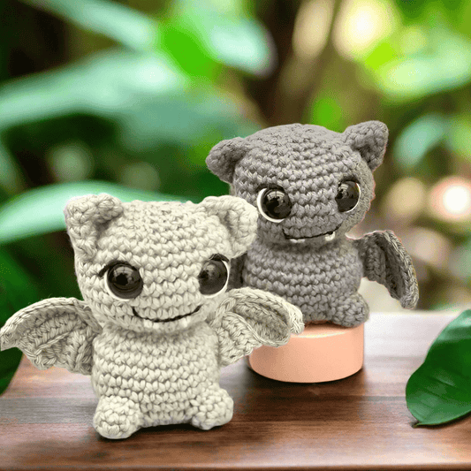 Two crocheted bat plushies, one white and one gray, sitting on a wooden surface with green leaves.